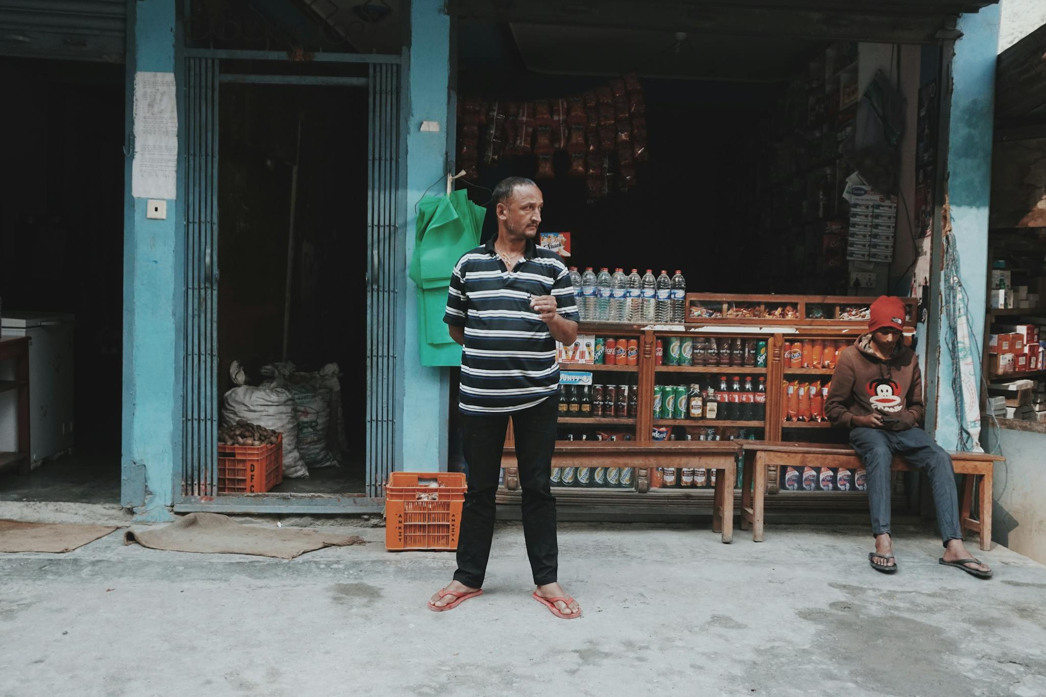 Two men outside a convenience store, one standing and one seated, with various products on display.